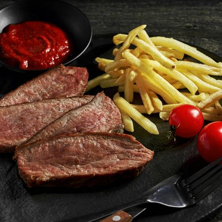 Sliced beef steak with French fries and cherry tomatoes on a black plate. Delicious American lunch with red wine.の写真素材