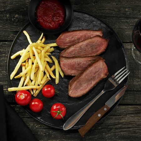 Delicious medium rare steak with French fries and fresh cherry tomatoes on a black plate. Classic meat meal with a glass of red wine. Top view, directly above shot.の写真素材