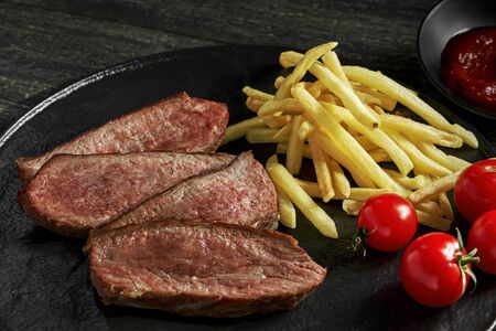 Steak with French fries, fresh cherry tomatoes and sauce on a rustic wooden table. Wholesome classic American lunch on a black plate. Closeup shot.の写真素材
