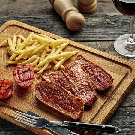 Steak with French fries and grilled cherry tomatoes on a cutting board. Classic meat meal with red wine on a wooden table with black tablecloth.の写真素材