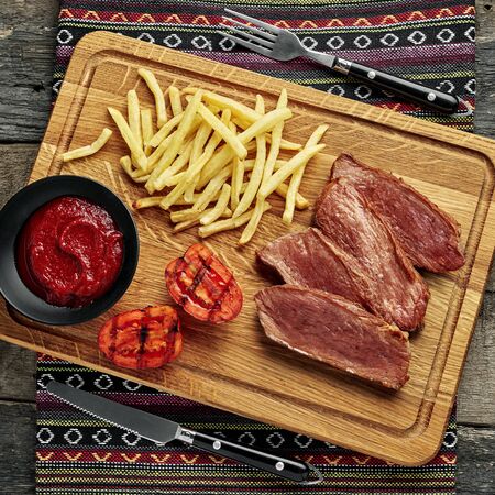 Medium rare sirloin steak with chips and grilled tomatoes on a rustic wooden table with ethnic tablecloth. Classic European lunch. Top view, directly above.の写真素材