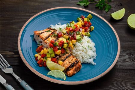 Grilled fillet of salmon with rice garnish and exotic fruit sauce made of mango, avocado, pepper and onion on a rustic wooden table.の写真素材