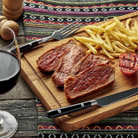 Steak with French fries and grilled cherry tomatoes on a cutting board. Classic meat meal with red wine on an ethnic tablecloth.の写真素材