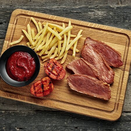 Steak with French fries, grilled cherry tomatoes, sauce and red wine on a wooden table. Wholesome American lunch served on a cutting board. Top view, directly above.の写真素材