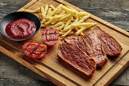 Steak with French fries, grilled cherry tomatoes, sauce and red wine on a rustic wooden table. Wholesome American lunch served on a cutting board. Close-up shot.の写真素材