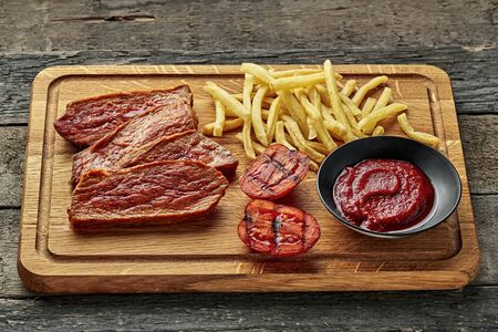 Steak with French fries, grilled cherry tomatoes, sauce and red wine on a rustic wooden table. Wholesome American lunch served on a cutting board.の写真素材