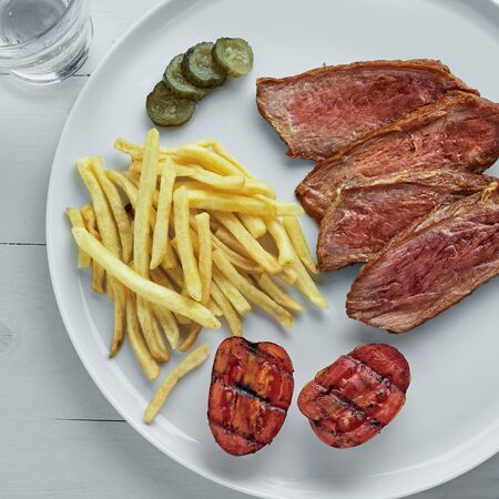 Sliced beef steak with French fries, pickled cucumber and grilled cherry tomatoes on a plate. Delicious American lunch with sparkling water. Top view, directly above.の写真素材