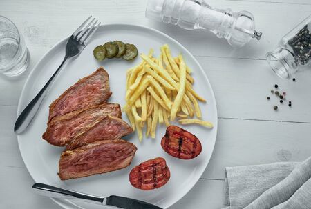 Sliced beef steak with French fries, pickled cucumber and grilled cherry tomatoes on a plate. Delicious American lunch with sparkling water. Top view, directly above.の写真素材