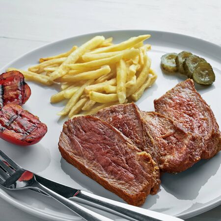 Medium rare sirloin steak with chips, pickled cucumber, grilled cherry  and a glass of sparkling water on a white wooden table with tablecloth. Classic European lunch.の写真素材