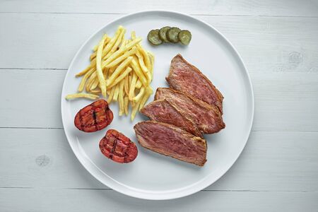 Gourmet medium rare beefsteak with French fries, pickled cucumber and grilled cherry tomatoes on a white plate. Top view, directly above shot.の写真素材