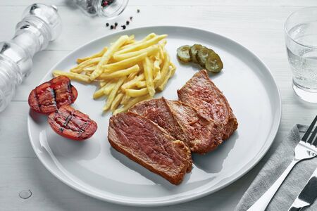 Sliced beef steak with French fries, pickled cucumber and grilled cherry tomatoes on a plate. Delicious American lunch with sparkling water.の写真素材
