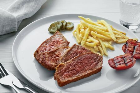 Medium rare sirloin steak with chips and a glass of sparkling water on a white wooden table with tablecloth. Classic European lunch.の写真素材