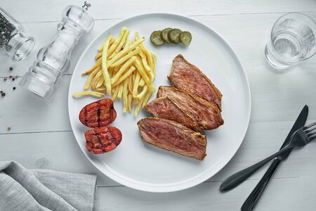 Steak with French fries and grilled cherry tomatoes on a plate. Classic meat meal with sparkling water on a white wooden table with a tablecloth. Top view, directly above.の写真素材