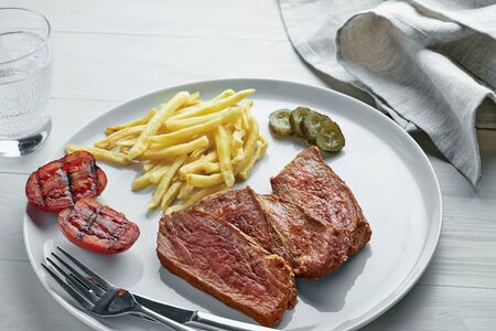 Steak with French fries, pickled cucumber, grilled cherry tomatoes and glass of sparkling water on a white wooden table. Wholesome American lunch.の写真素材