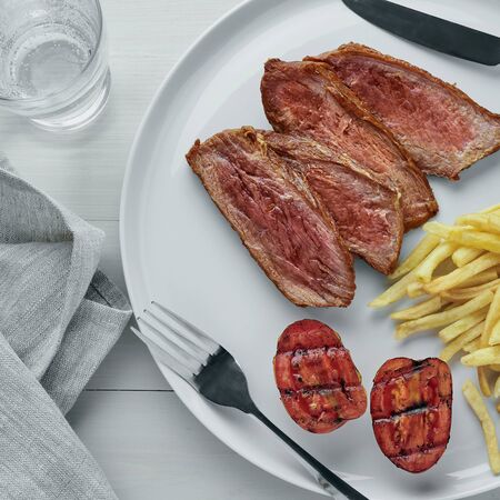 Steak with French fries, pickled cucumber, grilled cherry tomatoes and glass of sparkling water on a white wooden table. Wholesome American lunch. Top view, directly above.の写真素材