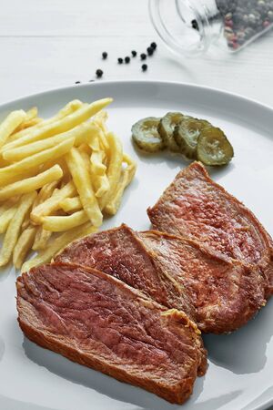 Gourmet medium rare beefsteak with French fries, pickled cucumber and grilled cherry tomatoes on a white plate with a glass of sparkling water.の写真素材