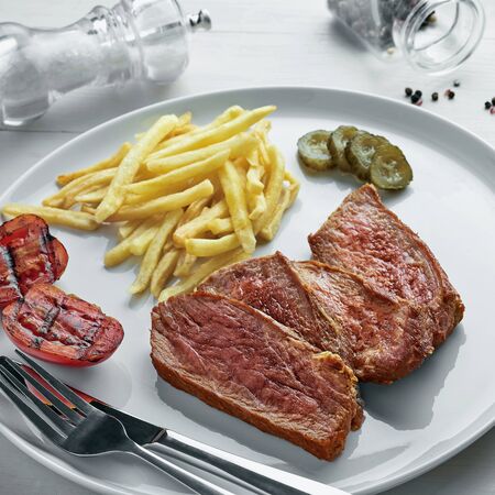 Steak with French fries, pickled cucumber, grilled cherry tomatoes and glass of sparkling water on a white wooden table. Wholesome American lunch.の写真素材