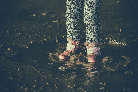 Little girl is playing in water, wet dirty shoes, in the middle of puddle, unforgettable moments, children's entertainments, tinting effect. Concept - child with dad on walk.の写真素材