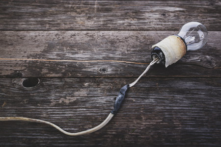 Concept - neglect of fire safety. Retro incandescent light bulb with damaged wires, on wooden background with space for textの写真素材