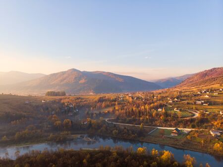 Beautiful autumn morning in picturesque Carpathians, Eastern Beskids. Soft sunlight awakens ancient village in valley between mountains, mountain river flows nearby.の写真素材