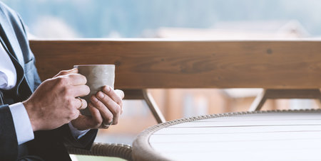 Businessman holding a hot tea cup in hands, on the terrace in the cafe. Work break, time of harmony and relaxation. Background in banner format, toning effect, copy spaceの写真素材