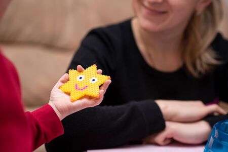 Little girl shows mom just crafted art toy from perler beads, beautiful smiled star. Fusible colorful beads. Toy that develops the imagination and creativity of child.の写真素材