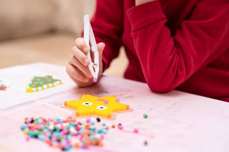 Little girl holds a tweezers with bead, creates art toy from fusible colorful beads, last touches. Also known as perler beads. Toy that develops the imagination of child. Process close-upの写真素材