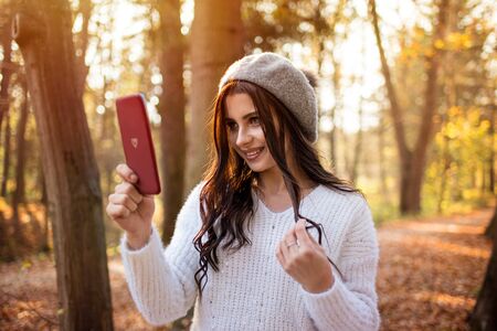 Young beautiful smiling caucasian woman taking selfie on cellphone in autumn park. Good sunny day and relaxation in nature creates a great moodの写真素材