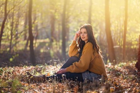 Young beautiful caucasian woman sits on the ground in a autumn park among high trees. Good sunny day it is great opportunity for relaxation and harmony with nature.の写真素材
