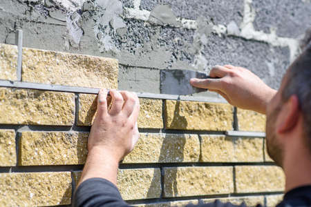 Man decorates the facade of the fence with decorative torn bricks, close up view. Work at home during quarantine, DIYの写真素材