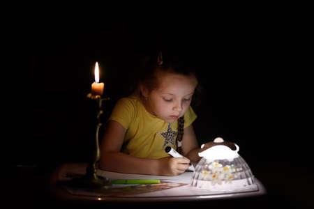Little girl draws fantasy characters near candle in a dark room. Child doing favorite thing during a power outage.の写真素材