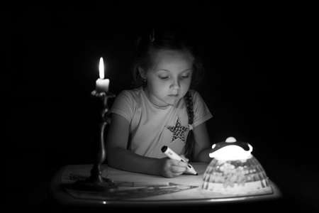 Little girl draws fantasy characters near candle in a dark room. Child doing favorite thing during a power outage. In black and white colors.の写真素材