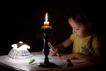 Little girl draws fantasy characters near candle in a dark room. Child doing favorite thing during a power outage.の写真素材