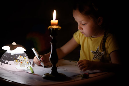 Little girl draws fantasy characters near candle in a dark room. Child doing favorite thing during a...