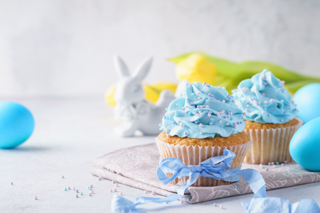 Homemade vanilla cupcakes decorated with sprinkles and blue ribbon. Bunny, colored eggs and yellow tulips in the background.の写真素材