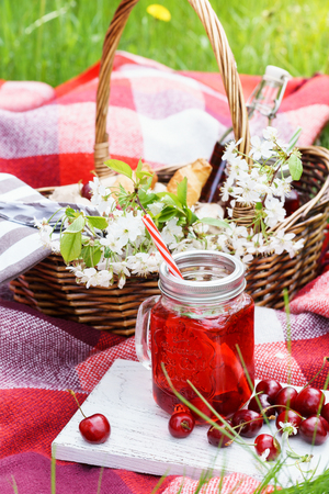 Cherry juice in mason jar and a picnic basket with food and flowers in the background. Outdoor picnic on grass.の写真素材