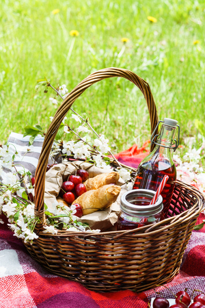 Picnic basket with food on green sunny lawn. Croissants, cherry berries and juice, flowers.の写真素材