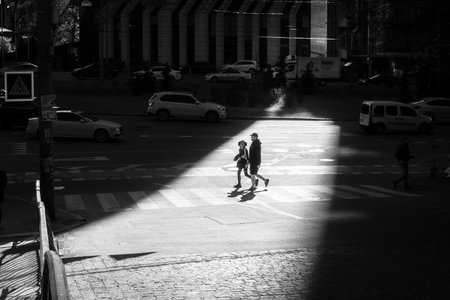 Editorial black and white image of a young couple walking across a pedestrian crosswalk in Kyiv, Ukraine, 2025.06.28. The composition features high-rise buildings, reflections architecture in glass, and strong contrast of light and shadow.のeditorial素材