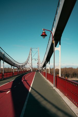 image of a modern suspension pedestrian and bicycle bridge with red railings and a two-tone path, stretching into the distance beneath a clear blue sky. The photo captures strong symmetry, clean architectural lines, and a sense of depth and openness.の写真素材