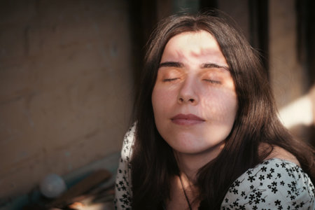 Natural portrait of a young brunette woman with closed eyes in warm sunlight. Soft shadow pattern across the face, calm mood, beauty and lifestyle concept in an intimate rustic outdoor setting.