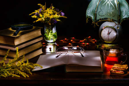 Flowers in a vase,a stack of books with a magnifying glass,an open book with glasses,a mug of tea and biscuits,table lamp with clock,berries rosehips.Illuminated by the flashlight.の写真素材