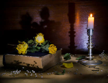 Gloomy still life.Consisting of three old yellow roses book candlestick with burning candle.の写真素材