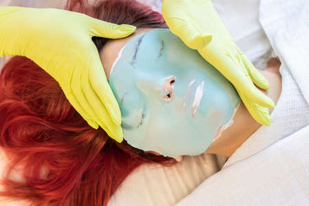 Hands beautician doctor put a face mask on the face of a beautiful young girl with red hair. Face care concept, lifestyle, beauty services, modern fashion, blog, notesの写真素材