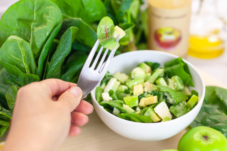Salad tasting: the cooks hand holds a fork with a spinach leaf, and a plate of fresh green vegetables and lettuce leaves is on the table with apples, spinach and vinegar on a light background. Farm organic product, home-made.の写真素材