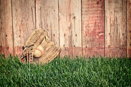 Old leather baseball mitt and ball on grass field against a rough wooden fenceの写真素材