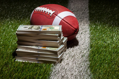 A football and a pile of one hundred dollar bills on a grass field with stripe and dark backgroundの写真素材