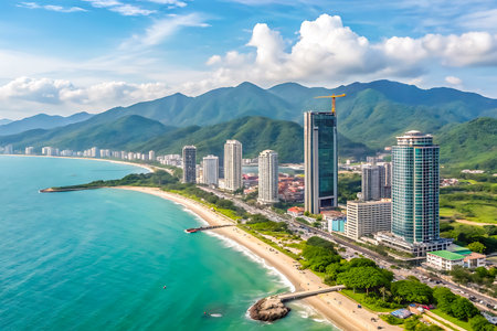 Turquoise ocean meets a sandy beach lined with modern skyscrapers and lush green mountains under a bright blue sky with white clouds.の素材