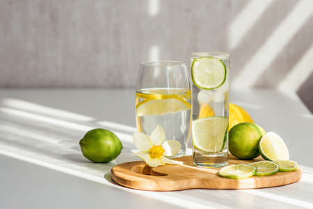 two glasses of water with lemon and lime on a wooden board and a flower of narcissus on a white tableの写真素材