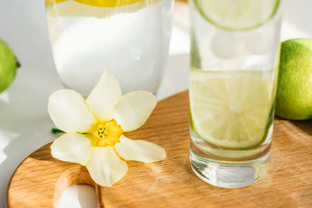 two glasses of water with lemon and lime on a wooden board and a flower of narcissus on a white tableの写真素材