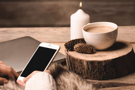 phone in the hands of the girl, a white cup with coffee, candle and cones on a wooden backgroundの写真素材